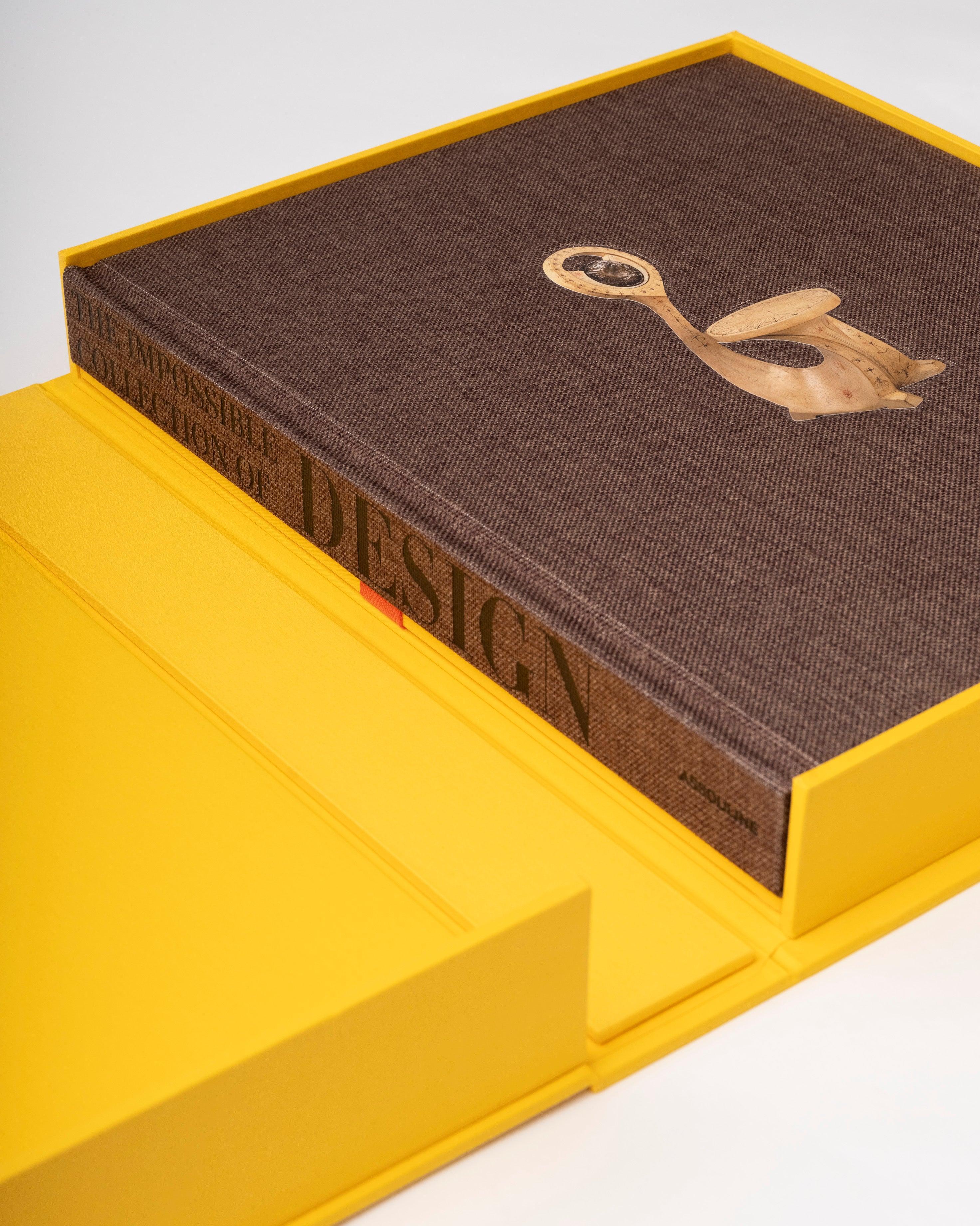 Libro Assouline de lujo en estuche amarillo, con tapa de tela marrón y silla de madera tallada.