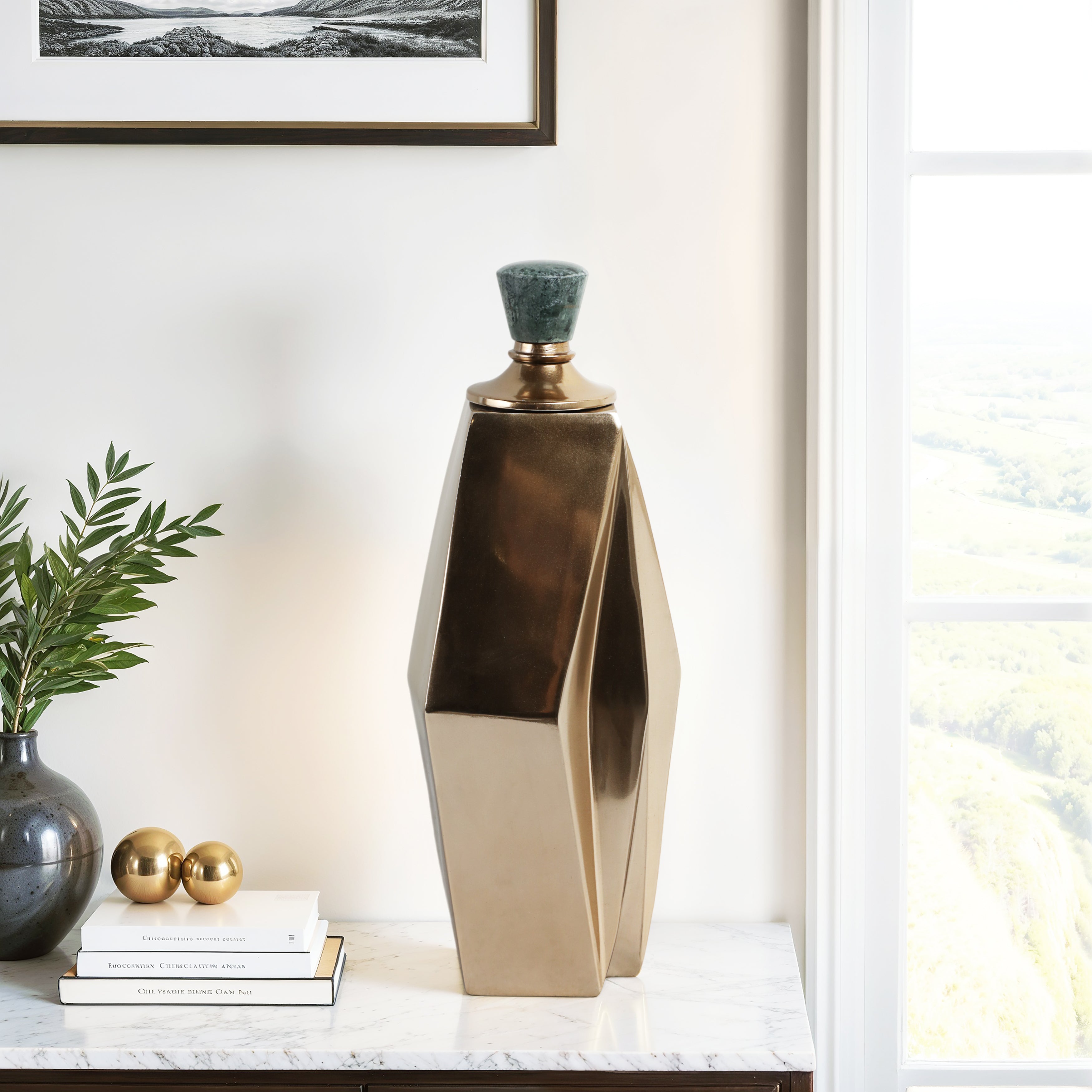 Botellón decorativo geométrico bronce sobre mesa de mármol con libros y planta verde.