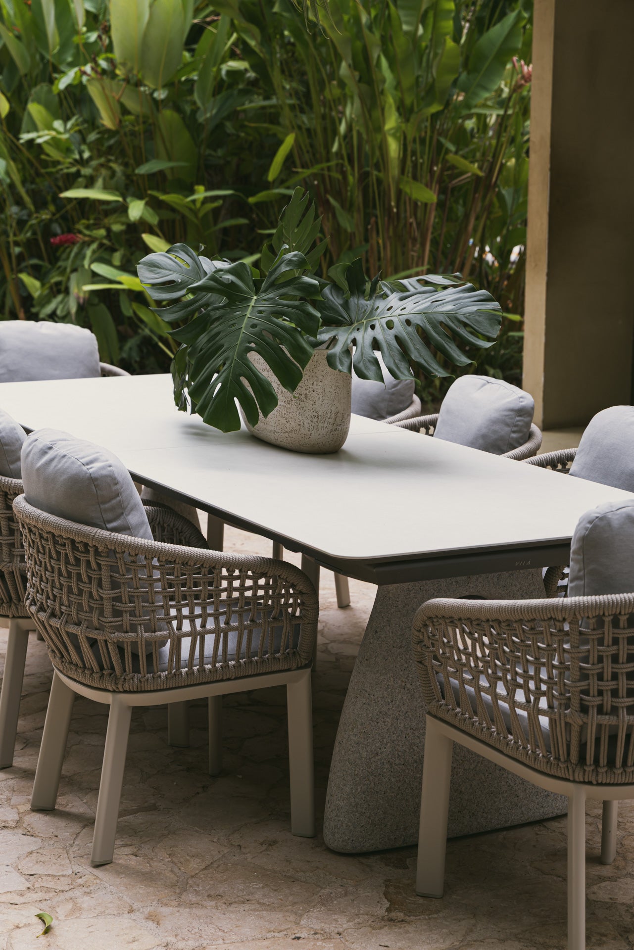 Mesa de comedor exterior de piedra sinterizada con sillas de ratán y plantas tropicales en un patio.