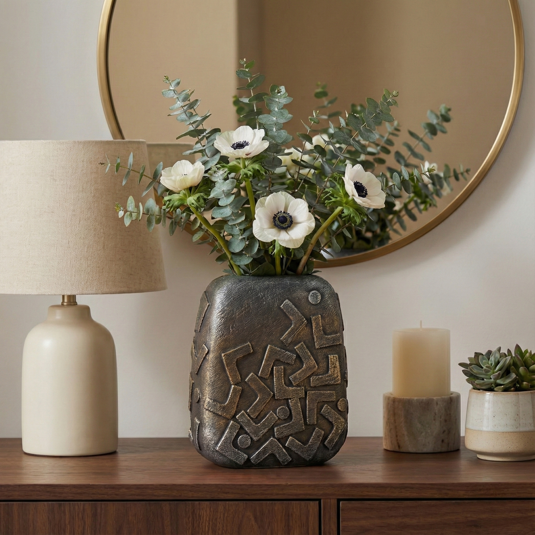 Jarrón decorativo bronce con flores blancas y eucalipto en mesa de madera, espejo y lámpara.