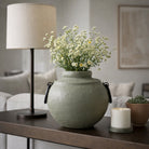 Jarrón esférico verde oliva con flores blancas sobre mesa de madera oscura en una sala de estar moderna y elegante.