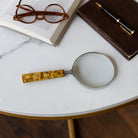 Lupa decorativa sobre mesa de mármol, junto a gafas, libro y pluma, en ambiente de lujo