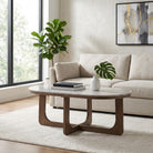 Mesa de centro ovalada de mármol y madera en sala de estar elegante con sofá beige y planta de interior.