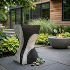 Mesa auxiliar escultural de concreto bicolor en patio exterior moderno con fogata y plantas.