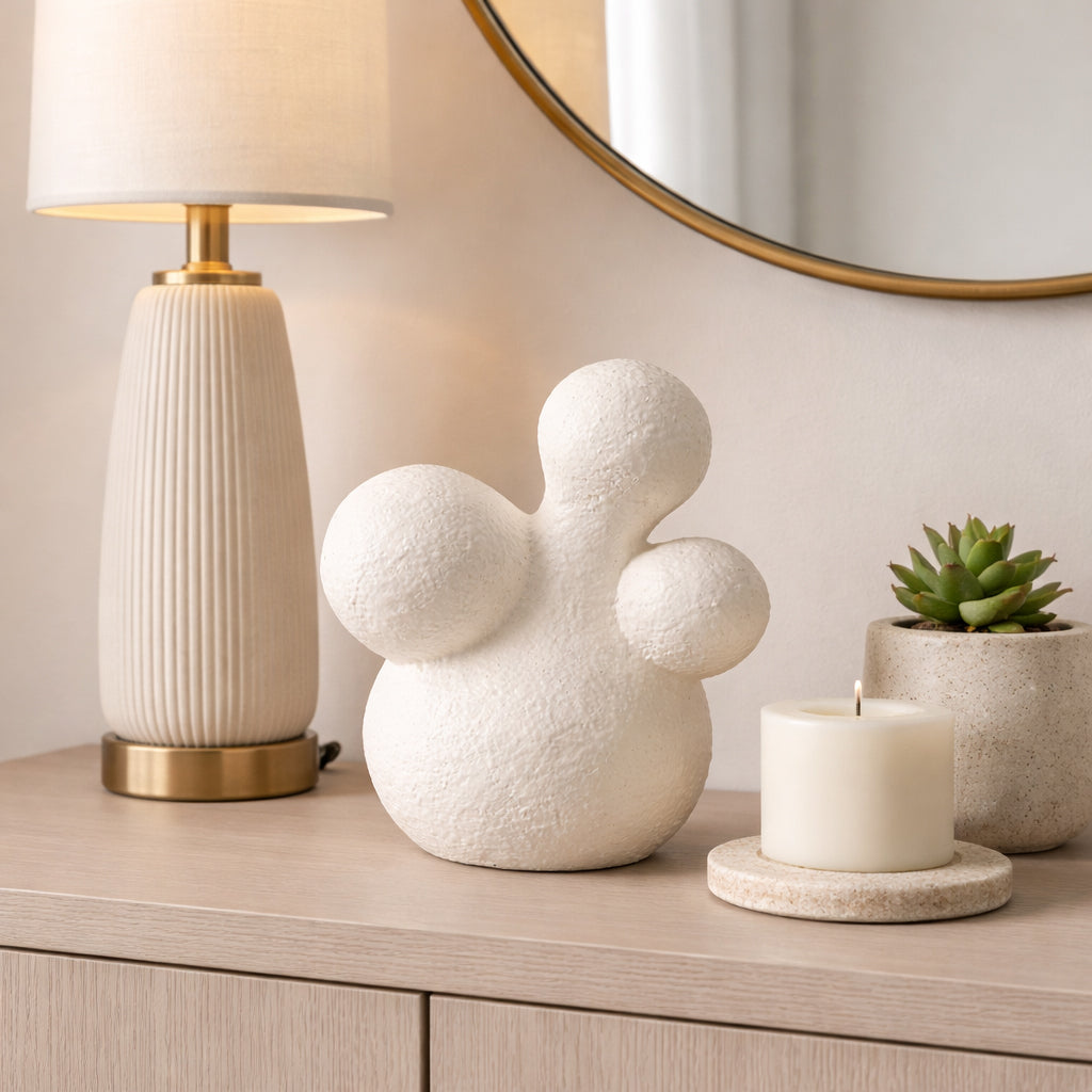 Escultura abstracta orgánica blanca cruda en mesa auxiliar con lámpara, espejo y planta decorativa.